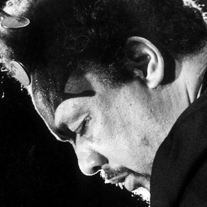 Close-up side profile of a man with a mustache leaning toward a guitar headstock, tuning pegs visible in black and white.