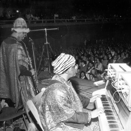 Two musicians perform on a crowded stage: a pianist in a shiny robe and zebra-striped turban at the keyboard, with a wind-instrument player beside him and a full audience in the background.