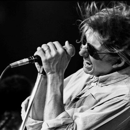 Black-and-white photo of a singer on stage, wearing sunglasses and gripping a microphone as their hair whipping in motion during a live performance,