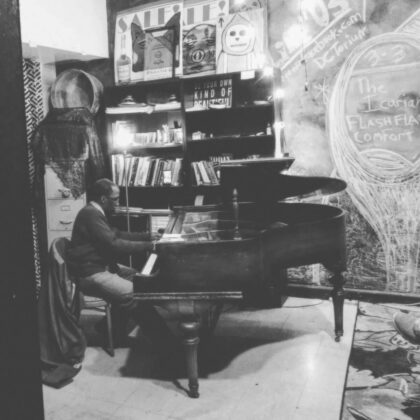 Man playing a grand piano in a small, cluttered room with bookshelves and posters on the wall