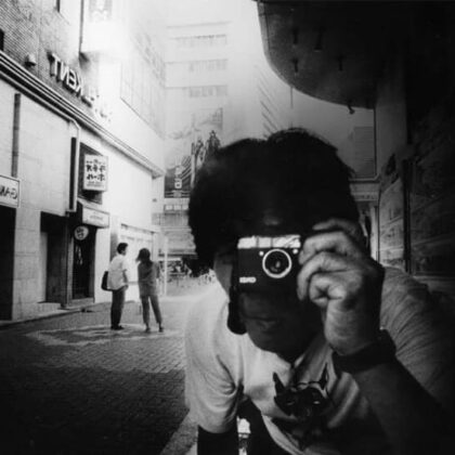 Black-and-white street photographer aiming a compact camera at the viewer in a city shopping district.