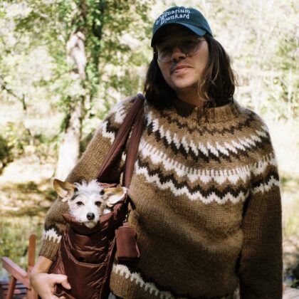 Person wearing a brown zigzag-knit sweater and teal cap, carrying a small white dog in a maroon sling bag outdoors in a sunlit forest.