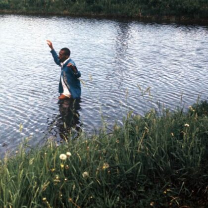 Man wading in a shallow river with grass and wildflowers in the foreground, arms raised as he stands in the water.