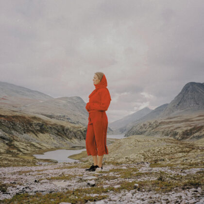 Person in a bright orange hooded jumpsuit stands on rocky terrain, gazing to the right at a mountainous landscape.