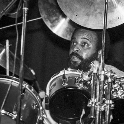 African American drummer with a beard playing a drum kit on stage, looking upward while performing on a dark stage.