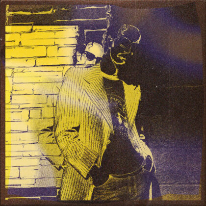 Two men in vintage suits leaning against a brick wall, posing together in a yellow-and-purple high-contrast photo treatment.