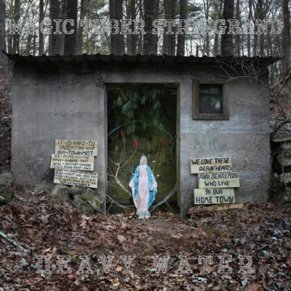 Album cover art: a concrete shack in a forest with a Mary statue in front and two handwritten signs, plus the title text 'MAGIC TUBER STRINGBAND' above and 'HEAVY WATER' below.