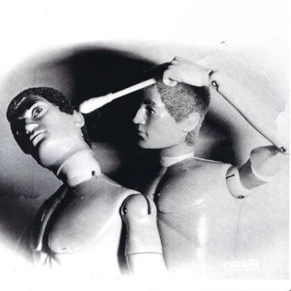 Two vintage jointed dolls in grayscale, with one doll holding a rod near the other’s head as if guiding or restraining it.