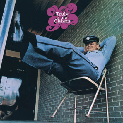 Promo image for Truly Fine Citizen showing a man in a vintage police hat lounging in a chair against a brick wall, smiling at camera.