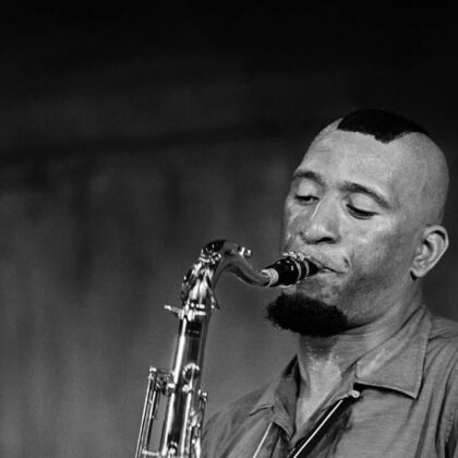 Black-and-white photo of a male saxophonist playing a curved sax, eyes closed, stage lighting in the background.