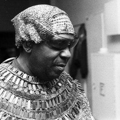 Man wearing an elaborate multi-row metallic necklace and headwrap, looking downward in a workshop or studio setting.
