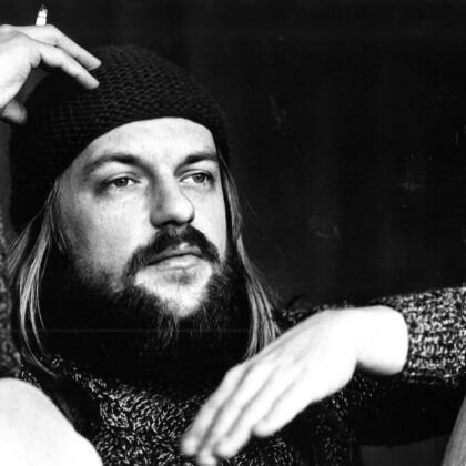 Black-and-white portrait of a bearded man wearing a knit beanie and textured sweater, resting his head on his hand with a cigarette nearby, gazing off to the side.