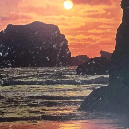 Sunset over a rocky coast with the sun low on the horizon and waves washing onto the shore.