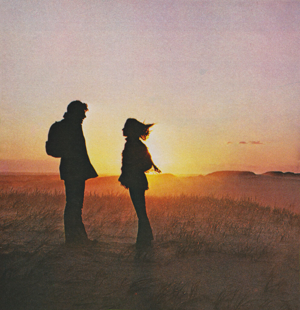 Silhouetted man and woman facing each other in a grassy field at sunset behind them.